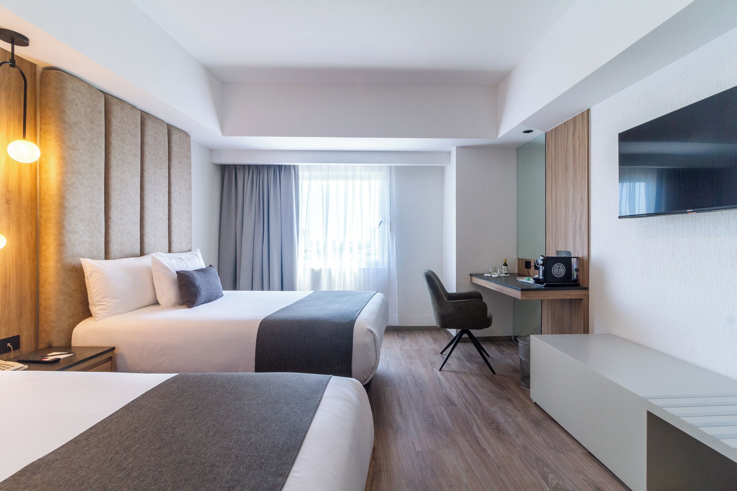 A hotel room with a bed, bedside table, lamp, curtains, a desk with a chair, and a television on the wall.