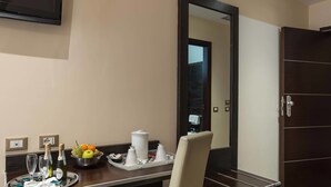 A hotel room with a wooden desk, a mirror, a bowl of fruit, and a tray with glasses and bottles. - Infinity Hotel San Pietro (Rome)