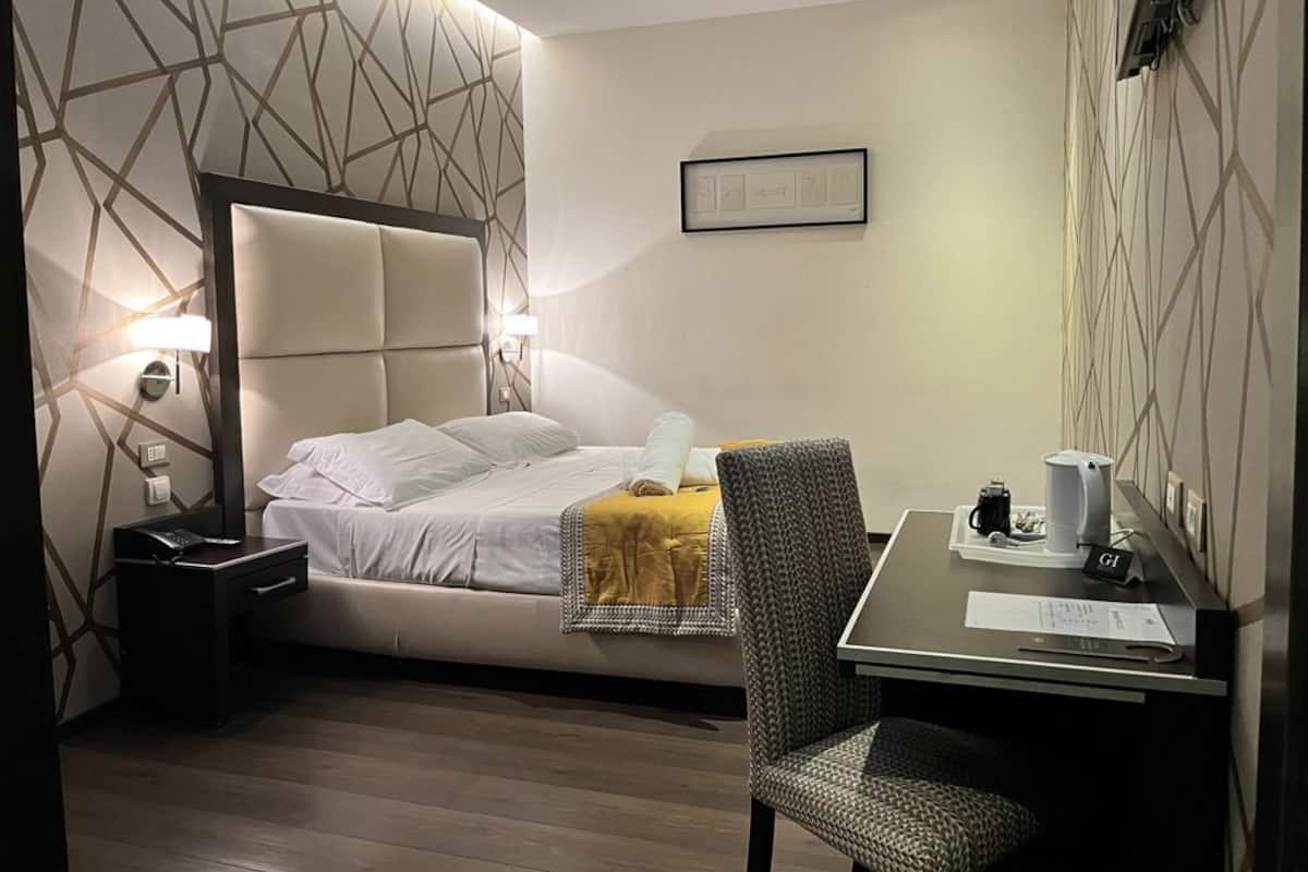 A modern hotel room with a large bed, a desk, and a chair. There is a framed picture on the wall and a lamp on the bedside table.