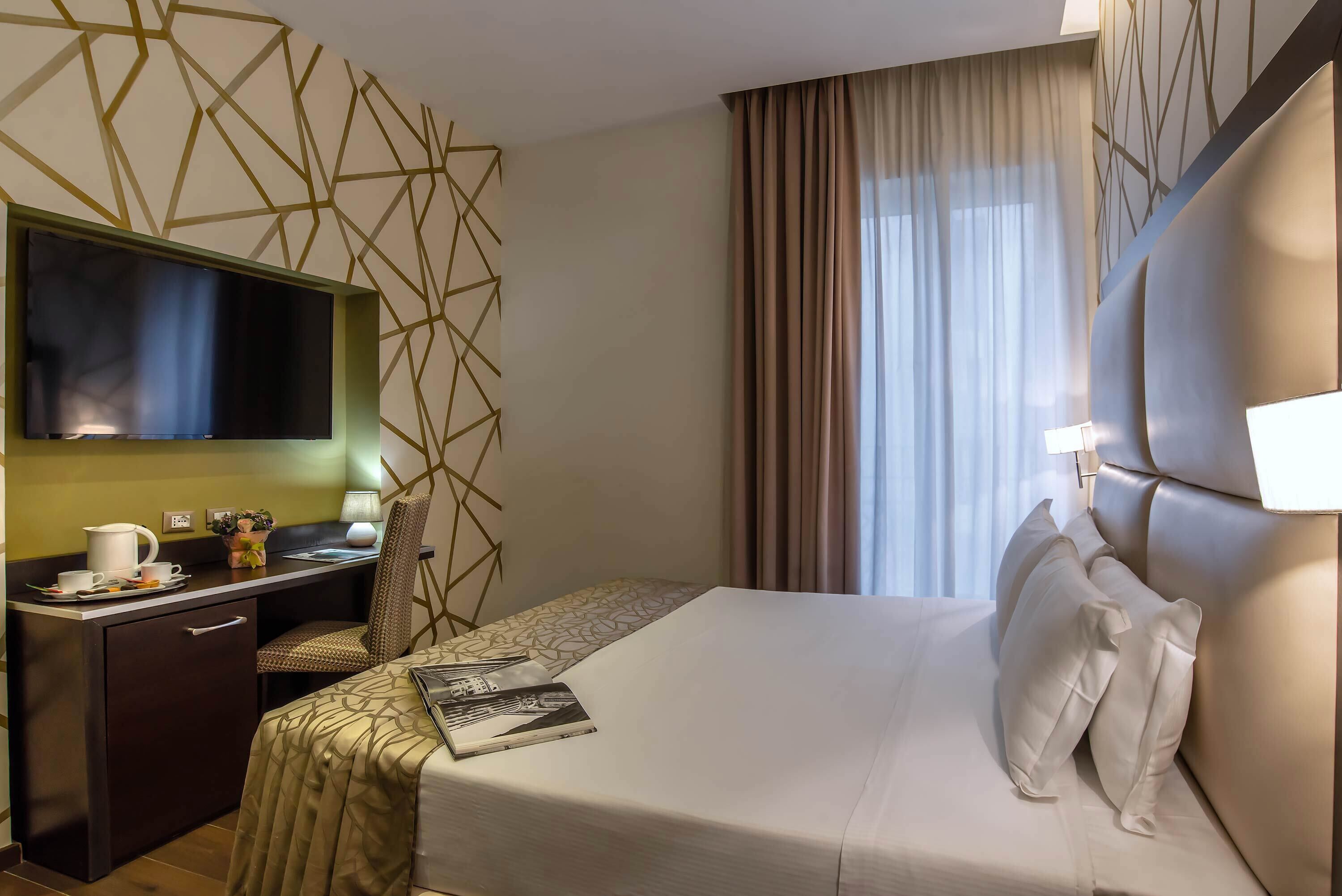 Quarto de hotel com cama, escrivaninha, cadeira, televisão e janela com cortinas.