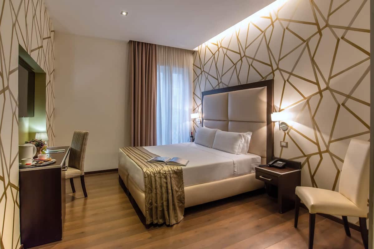A hotel room with a large bed, a desk with a chair, a bedside table with a lamp, and a chair by the window.