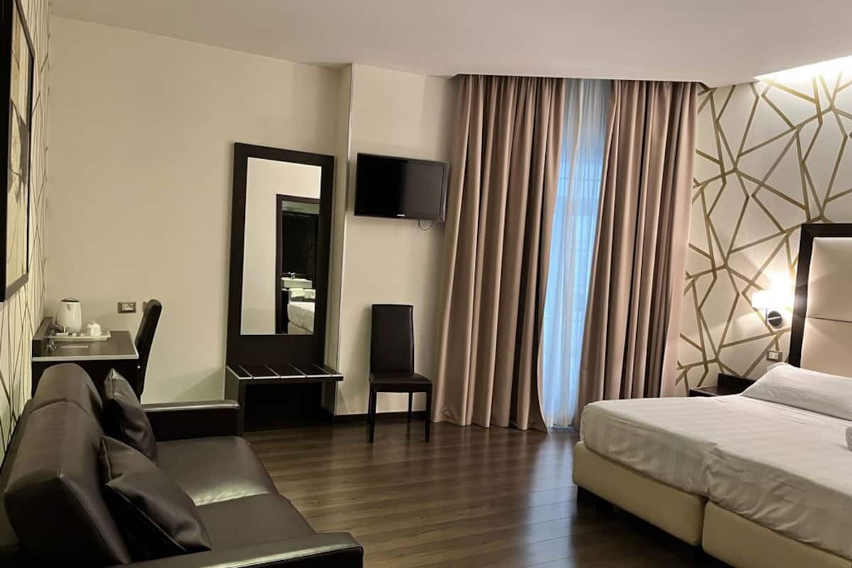 A hotel room with a bed, a sofa, a chair, a desk, a TV, and a window with curtains.