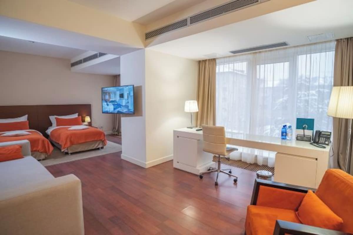 A hotel room with two beds, a desk with a chair, a TV, and a large window with curtains.