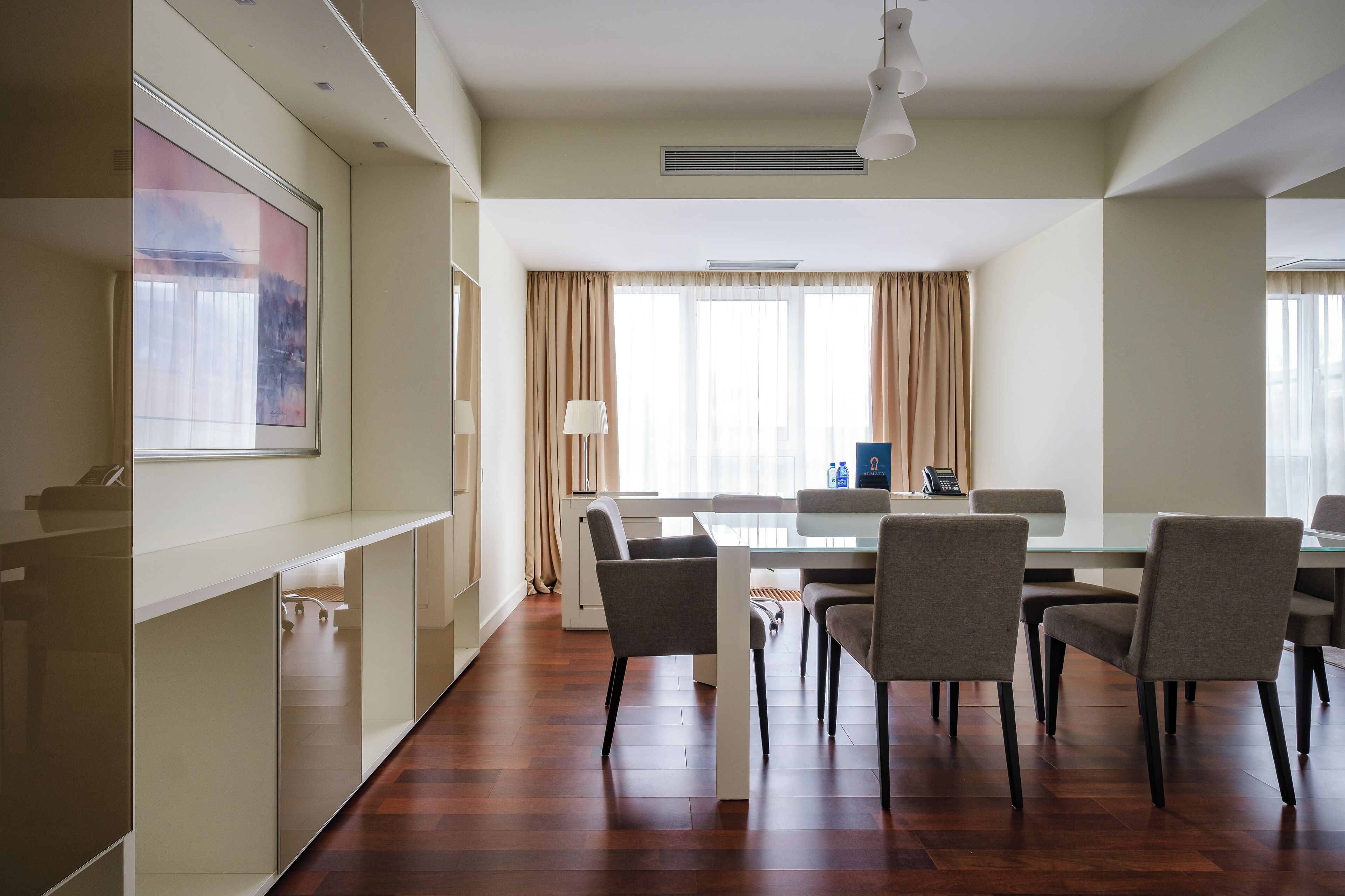 Quarto de hotel moderno com área de refeições, piso de madeira e grandes janelas com cortinas.