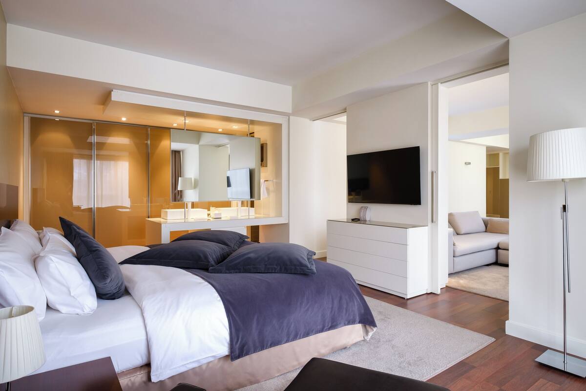 A modern bedroom with a large bed, a bedside table, a dresser, a TV, and a mirror.