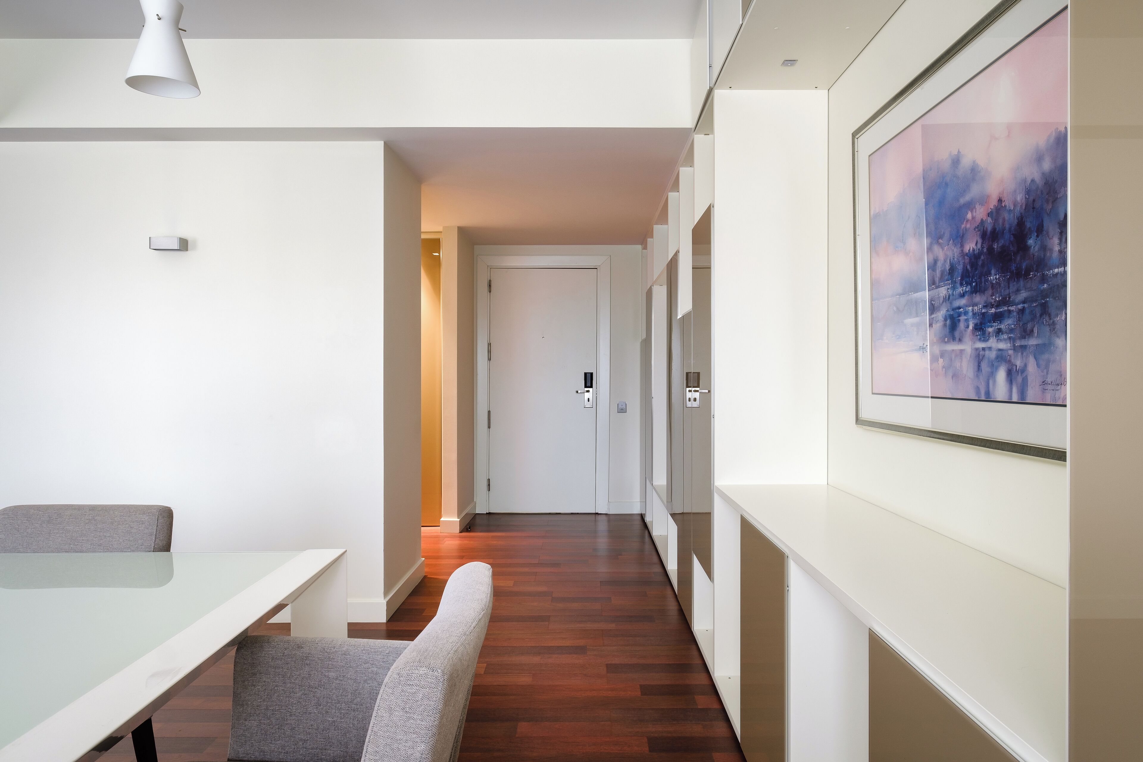 Een moderne hotelkamer met een glazen tafel, grijs gestoffeerde stoelen, een houten vloer, een witte deur en een lijstwerk schilderij aan de muur.