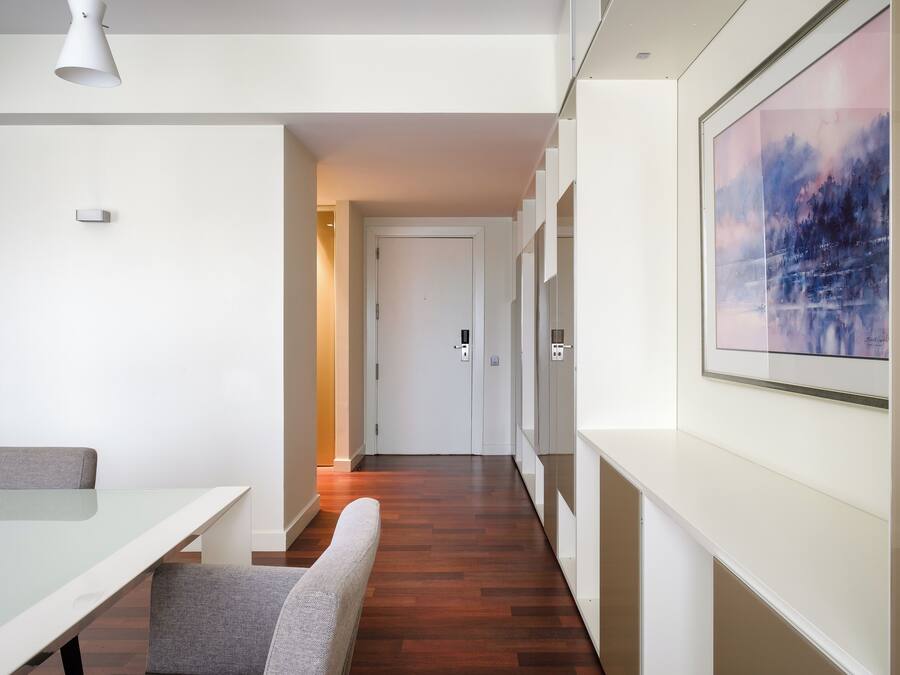 Een moderne hotelkamer met een glazen tafel, grijs gestoffeerde stoelen, een houten vloer, een witte deur en een lijstwerk schilderij aan de muur.