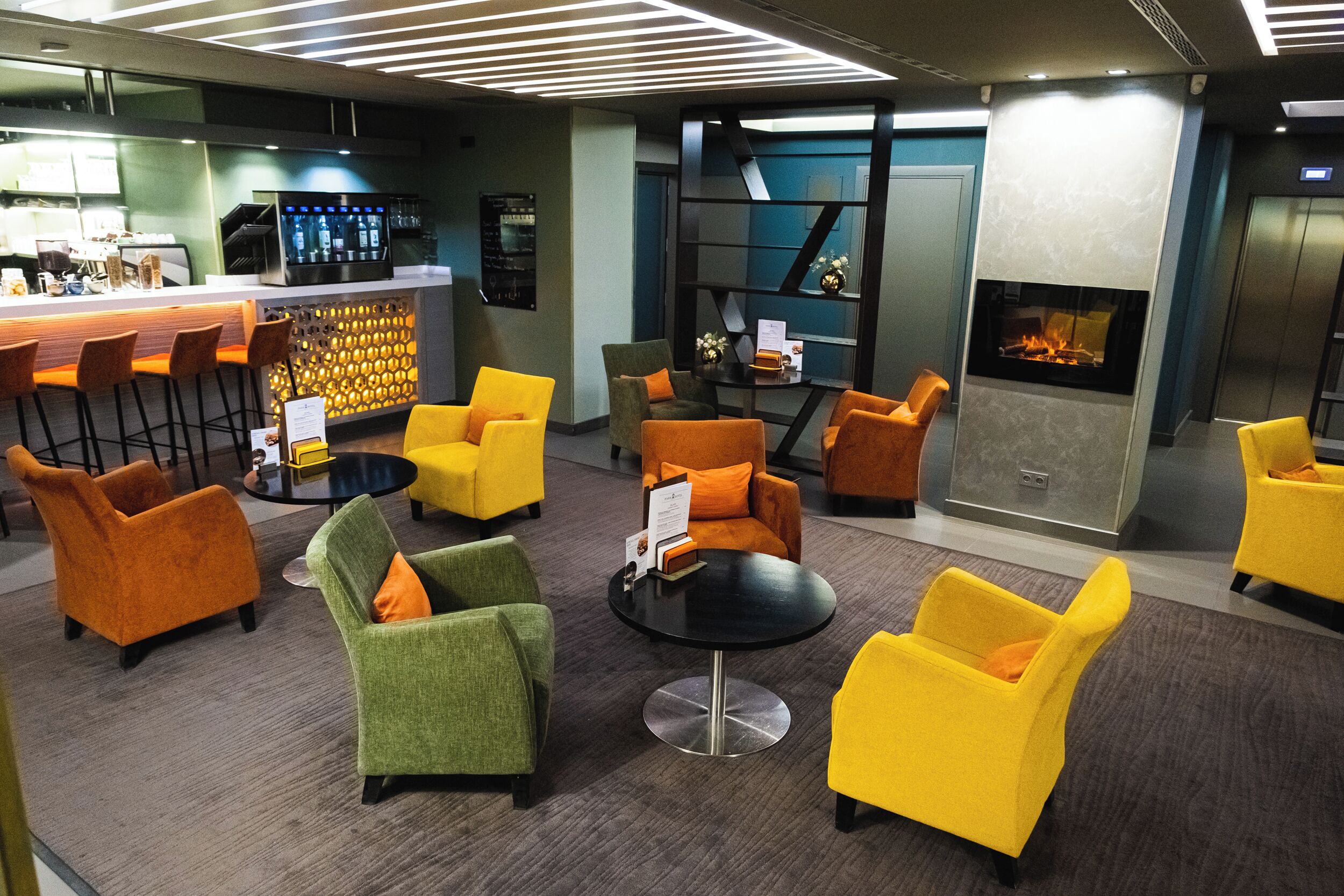 A modern lounge area with yellow and green chairs, a bar with a digital screen, and a fireplace.