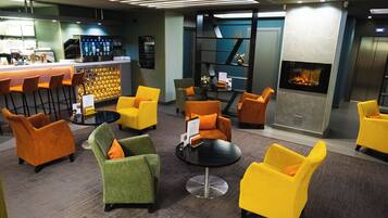 A modern lounge area with yellow and green chairs, a bar with a digital screen, and a fireplace.