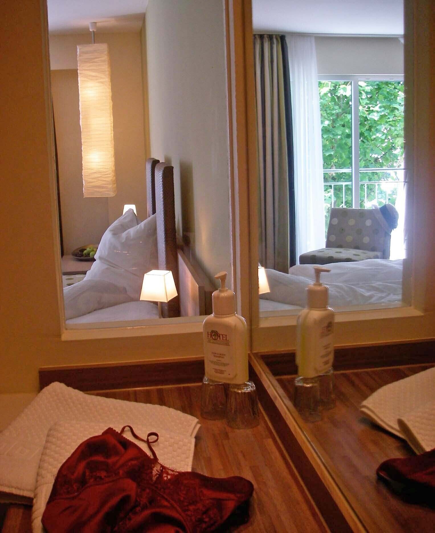 A hotel room with a large mirror, a bed with white linens, a bedside table with toiletries, and a view of greenery outside the window.