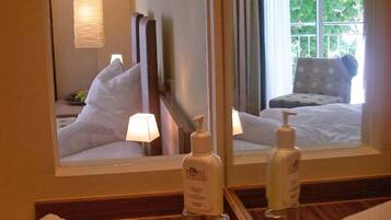 A hotel room with a large mirror, a bed with white linens, a bedside table with toiletries, and a view of greenery outside the window.