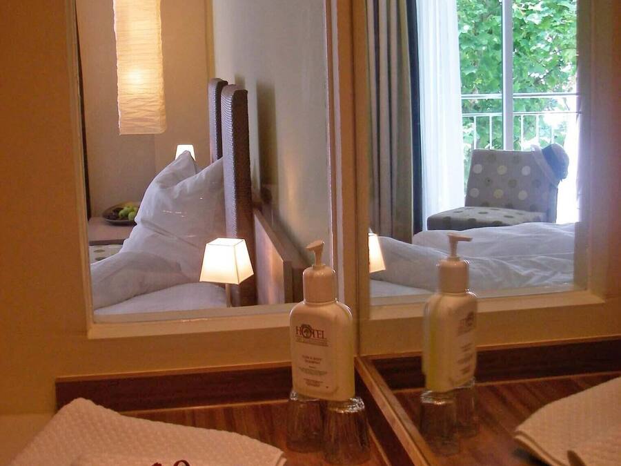 A hotel room with a large mirror, a bed with white linens, a bedside table with toiletries, and a view of greenery outside the window.