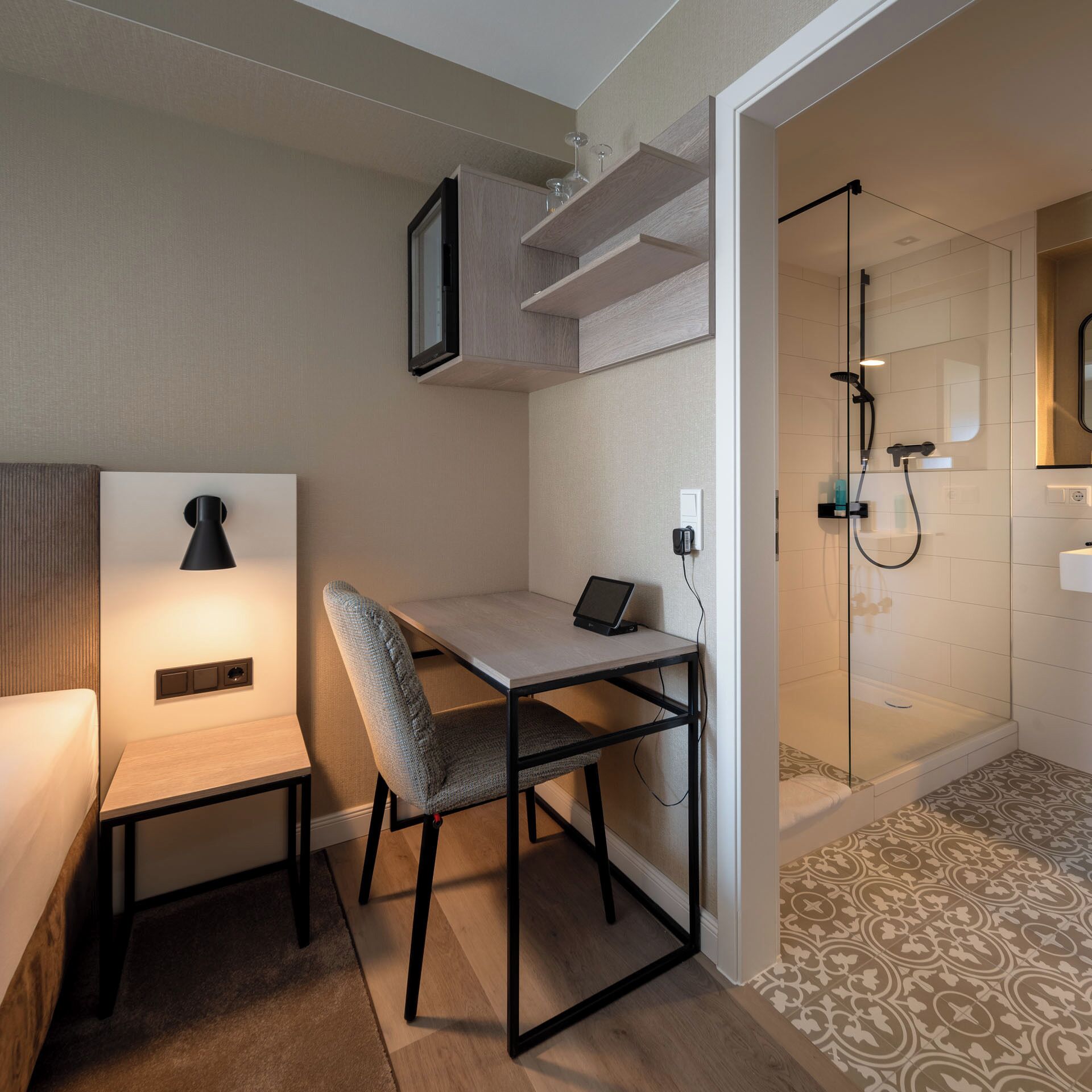A compact room with a bed, a desk with a chair, a small table, a wall-mounted TV, and a shower area.