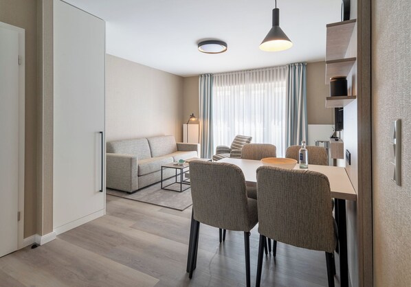 Una habitación de hotel moderna con sofá, mesa de comedor y sillas. Cuenta con un ventanal con cortinas, una lámpara de techo y un televisor sobre una estantería.