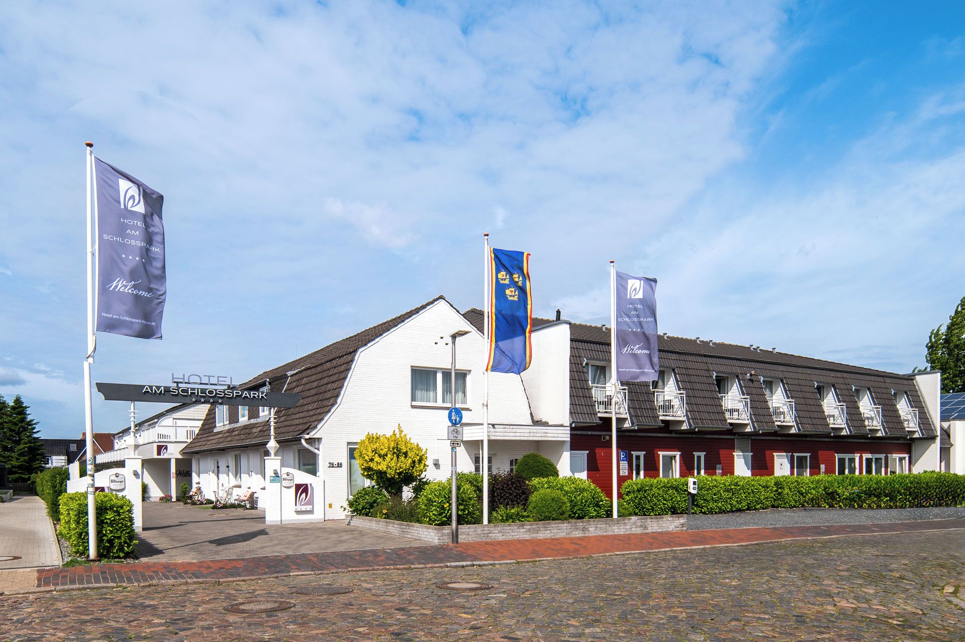 Et hotel med hvid facade, flere flag og en blå himmel.