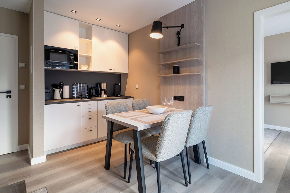 A modern kitchen with white cabinets, a microwave, and a coffee maker. There is a dining table with chairs and a TV mounted on the wall.