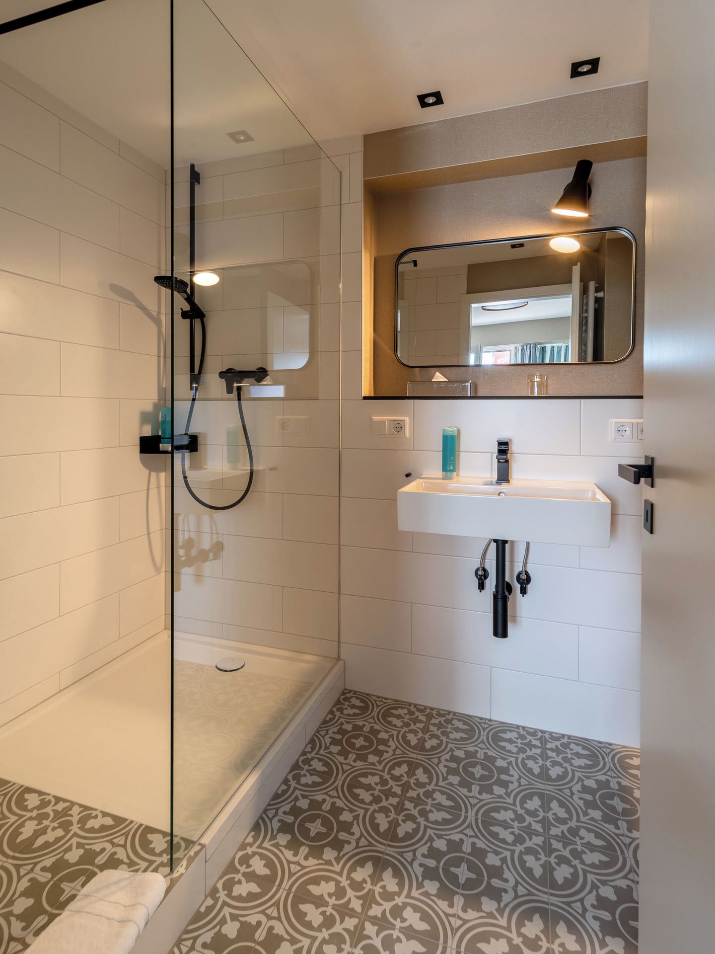 Un baño moderno con cabina de ducha de vidrio, un lavabo blanco con espejo superior y baldosas de suelo con diseño.