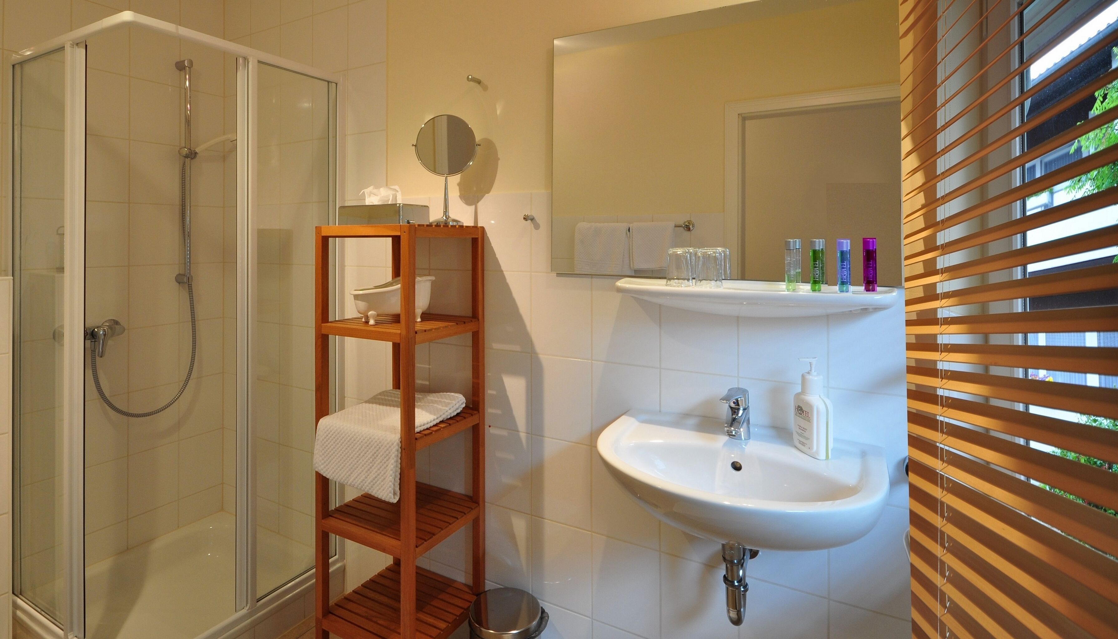 A bathroom with a glass-enclosed shower, a wooden shelf with toiletries, a sink, and a mirror.