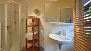 A bathroom with a glass-enclosed shower, a wooden shelf with toiletries, a sink, and a mirror.