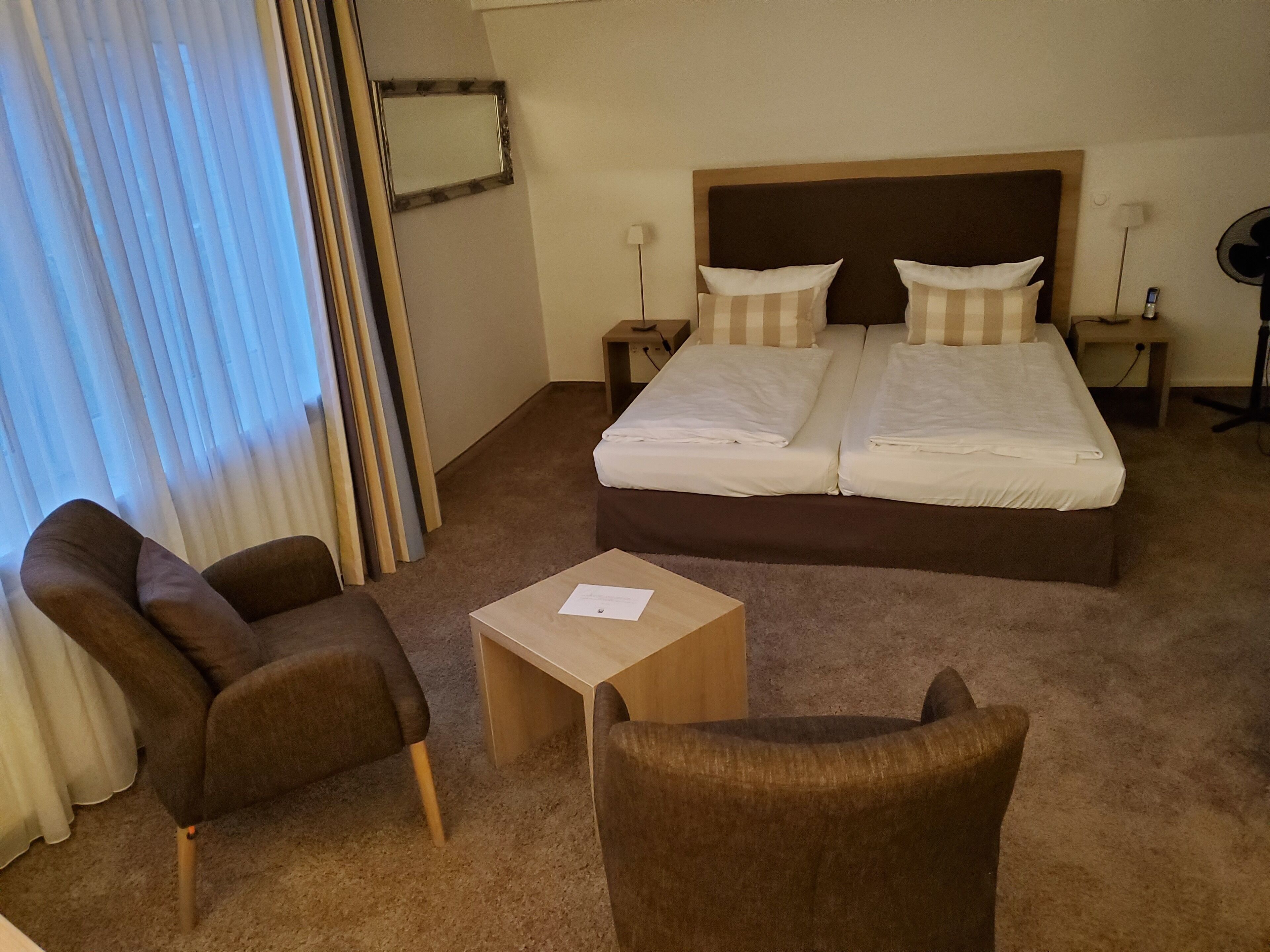 Quarto de hotel com cama de casal, duas mesas de cabeceira, uma cadeira, uma mesinha e janela com cortinas.