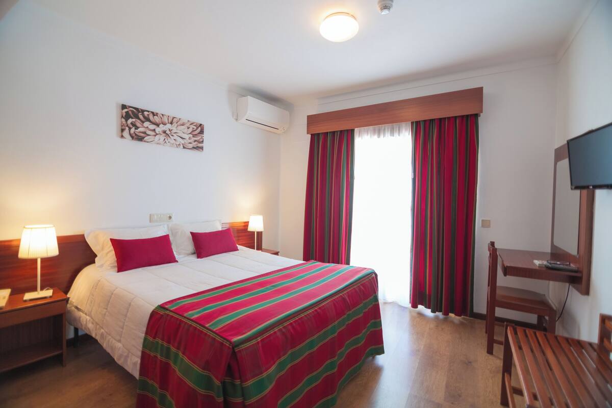 A hotel room with a bed, a desk, a chair, an air conditioning unit, a television, and a window with curtains.