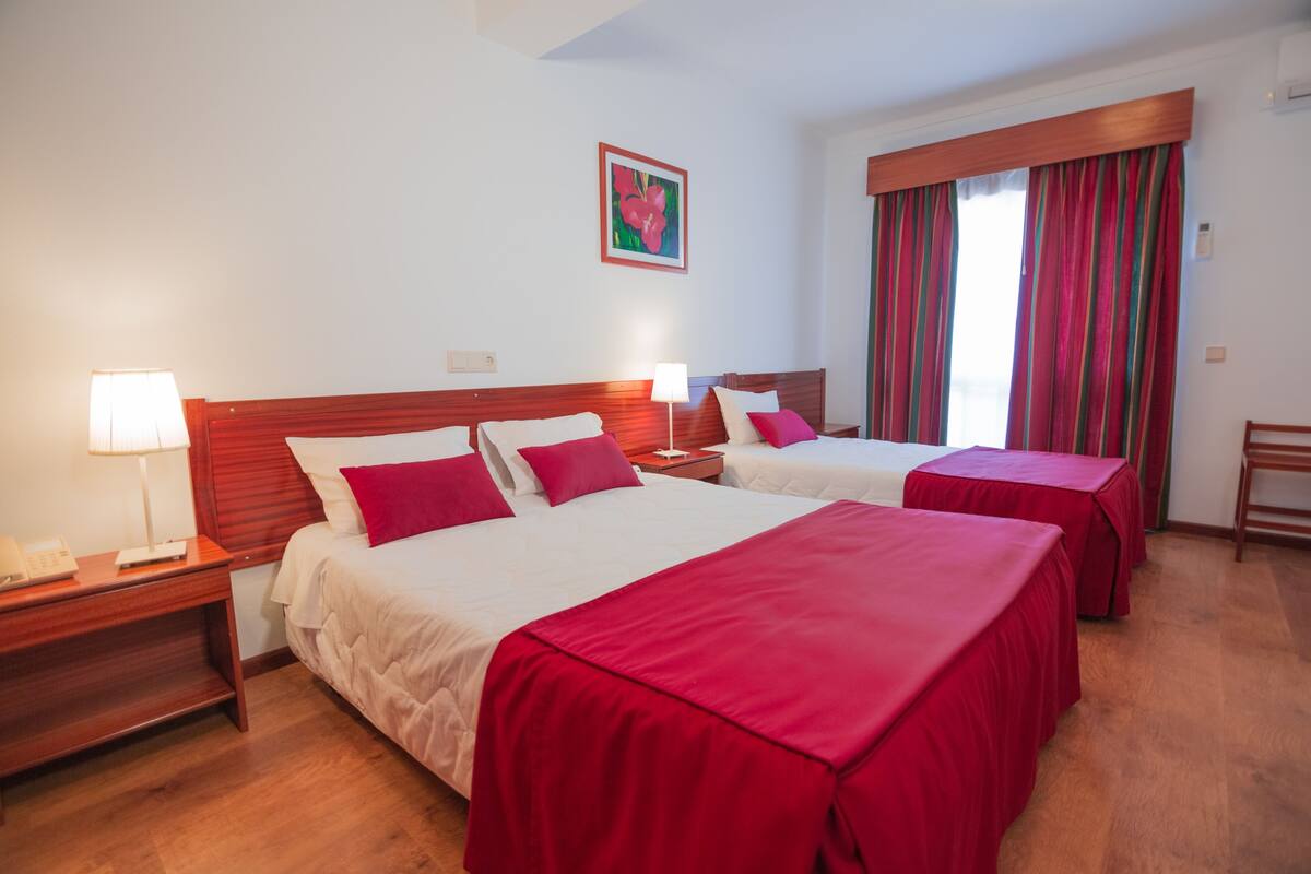 A hotel room with two beds, wooden headboards, and red bedspreads. There is a nightstand with a lamp and a telephone on each side of the beds. A window with striped curtains is visible in the background.