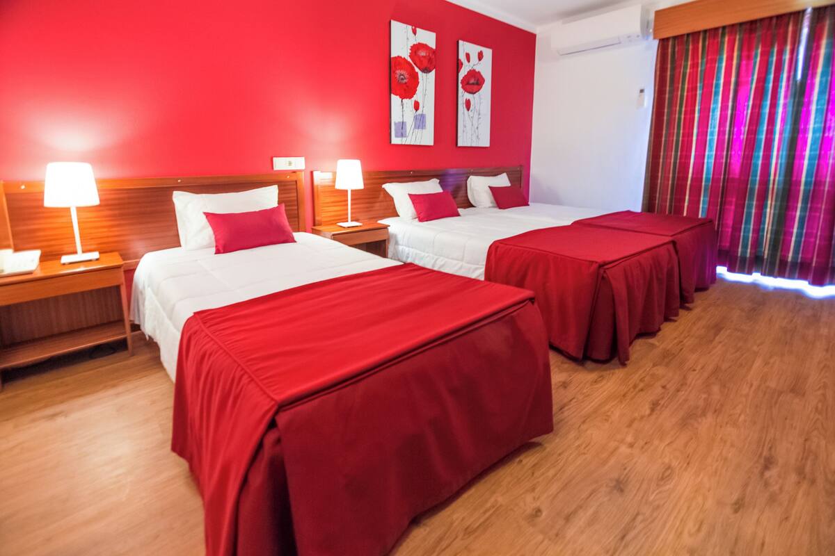 A hotel room with two beds, red bedding, wooden headboards, a wooden floor, and a red curtain.