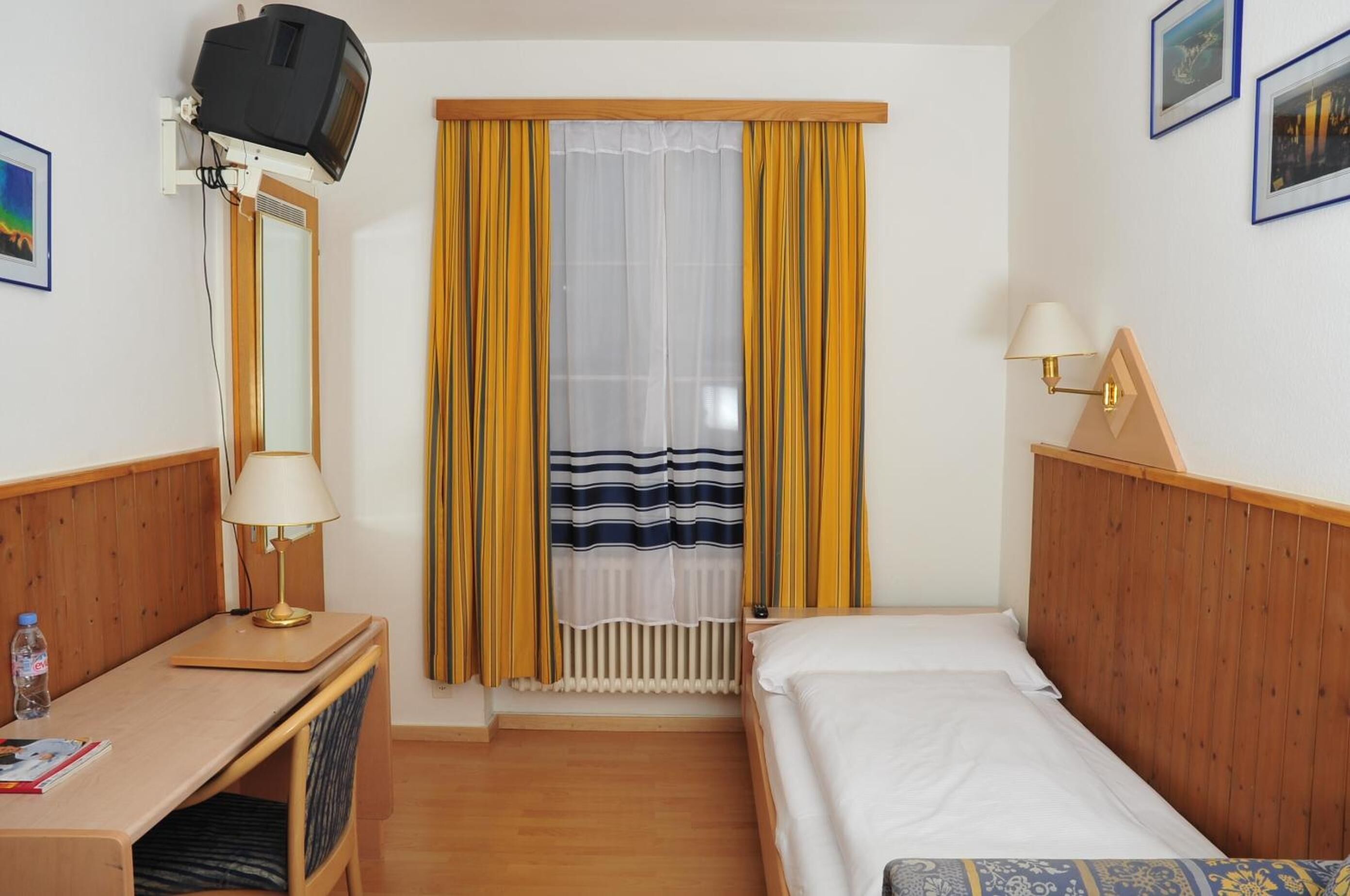 Quarto de hotel com cabeceira de madeira, uma cama, uma escrivaninha com abajur, televisão fixada na parede e uma janela com cortinas.
