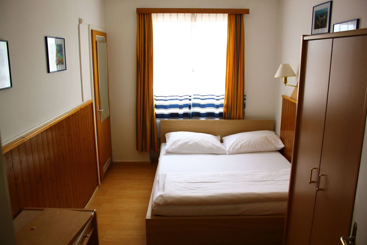 A hotel room with a bed, a wooden wardrobe, a desk, a window with curtains, and a view of the sea.
