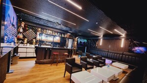 A modern bar area with a wooden floor, bar stools, and a counter with various bottles. There are seating areas with tables and chairs.