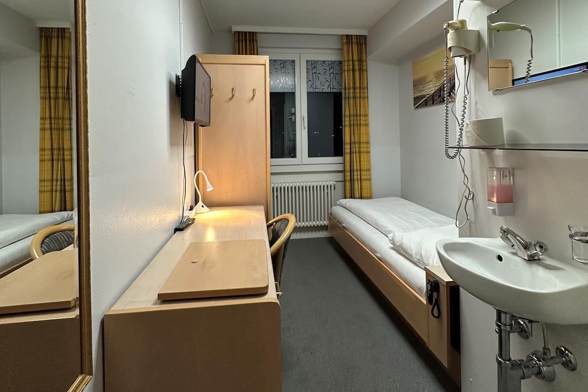 A compact hotel room with a single bed, a bedside table with a lamp, a small desk with a chair, a mirror, a window with curtains, and a bathroom with a sink and mirror.