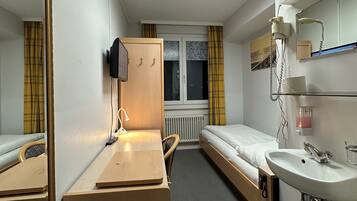 Une chambre d’hôtel de taille réduite, comprenant un lit simple, une table de chevet avec une lampe, un petit bureau avec une chaise, un miroir, une fenêtre avec des rideaux et une salle de bain avec un lavabo et un miroir.