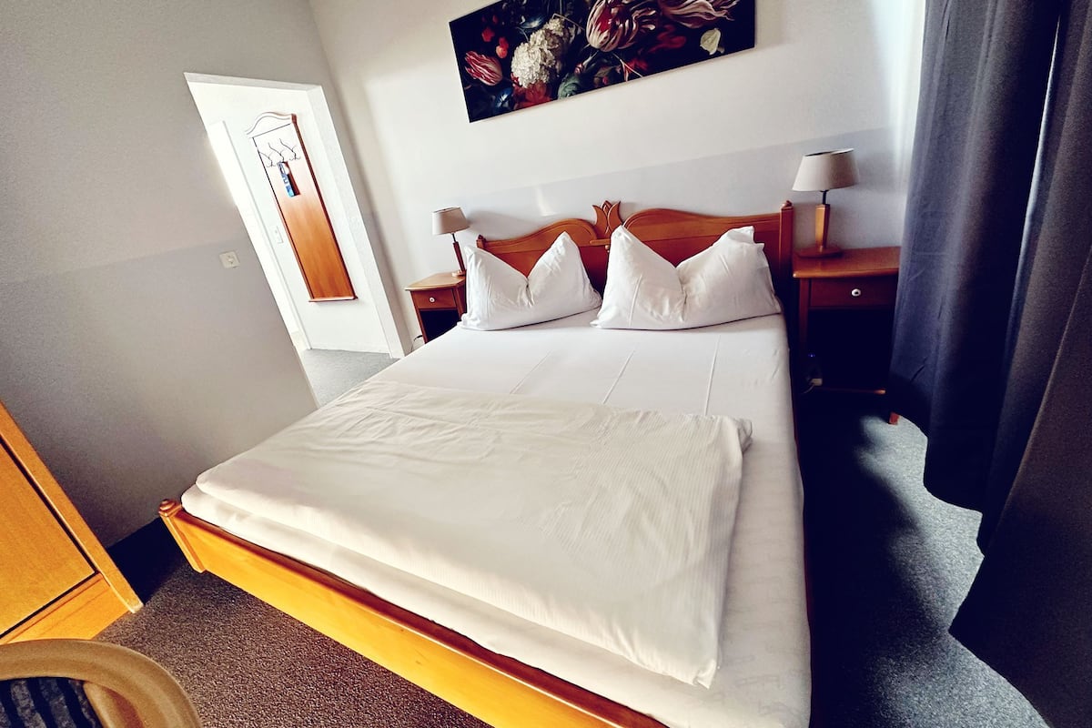 A hotel room with a large bed, wooden headboard, white bedding, a painting on the wall, a nightstand with a lamp, and a door.