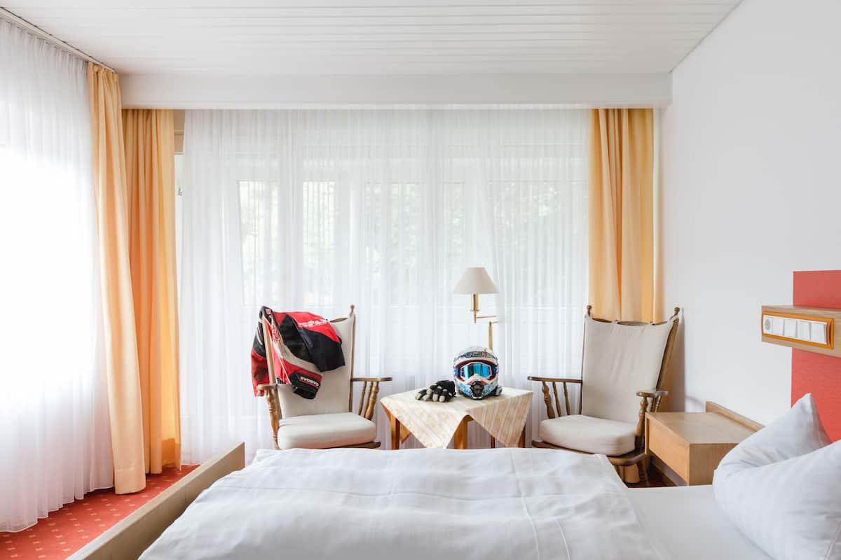 A hotel room with a bed, two armchairs, a small table, and a window with sheer curtains.