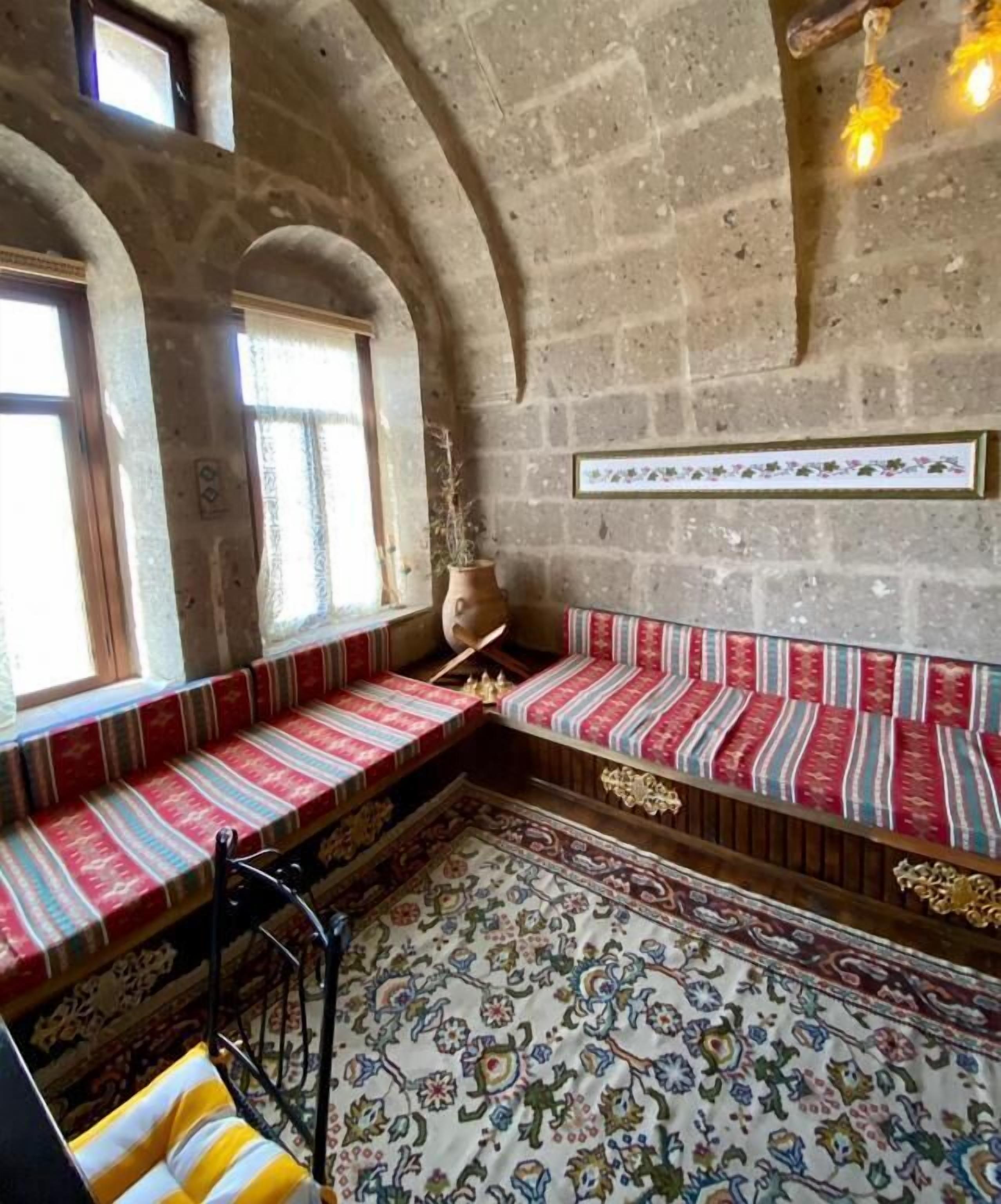 Een kamer met een stenen muur, een bank met rood-witte kussens, een kamerkleed met een patroon en een vaas aan de muur.