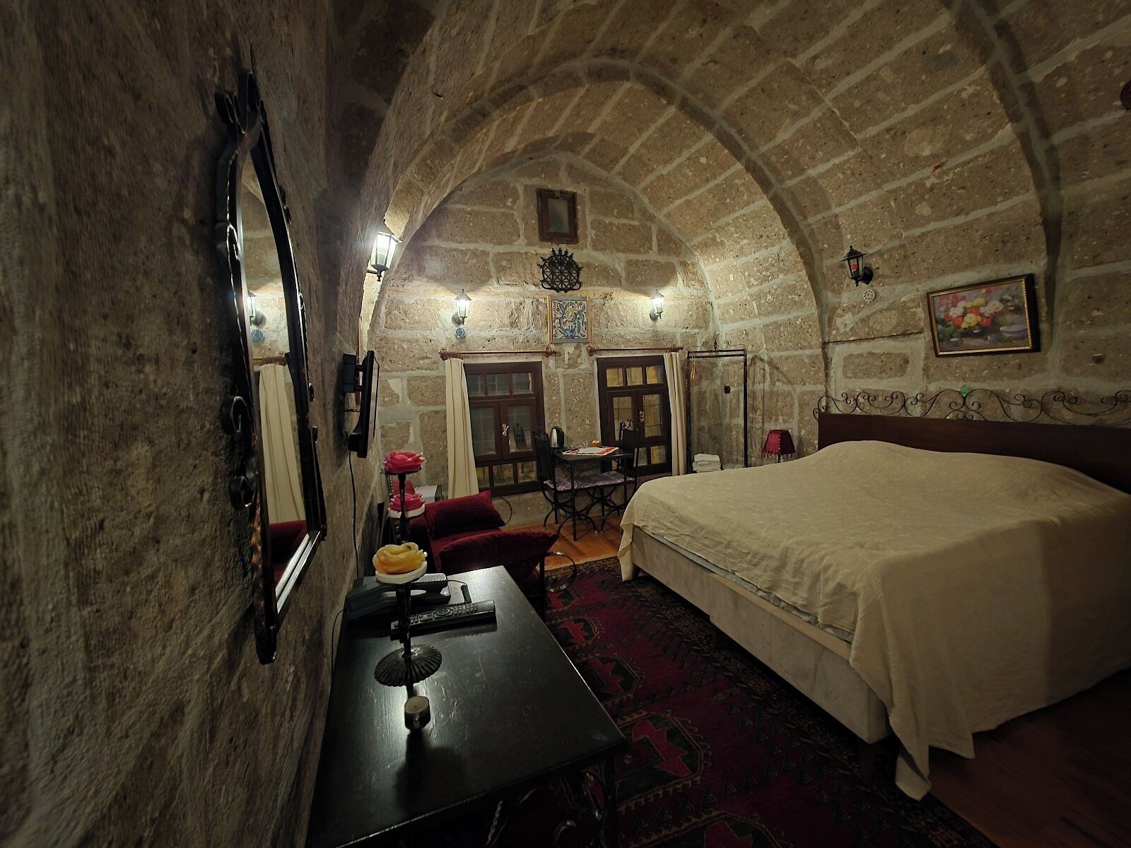 A room with a stone ceiling, a bed, a desk with a chair, and a small table with a lamp.
