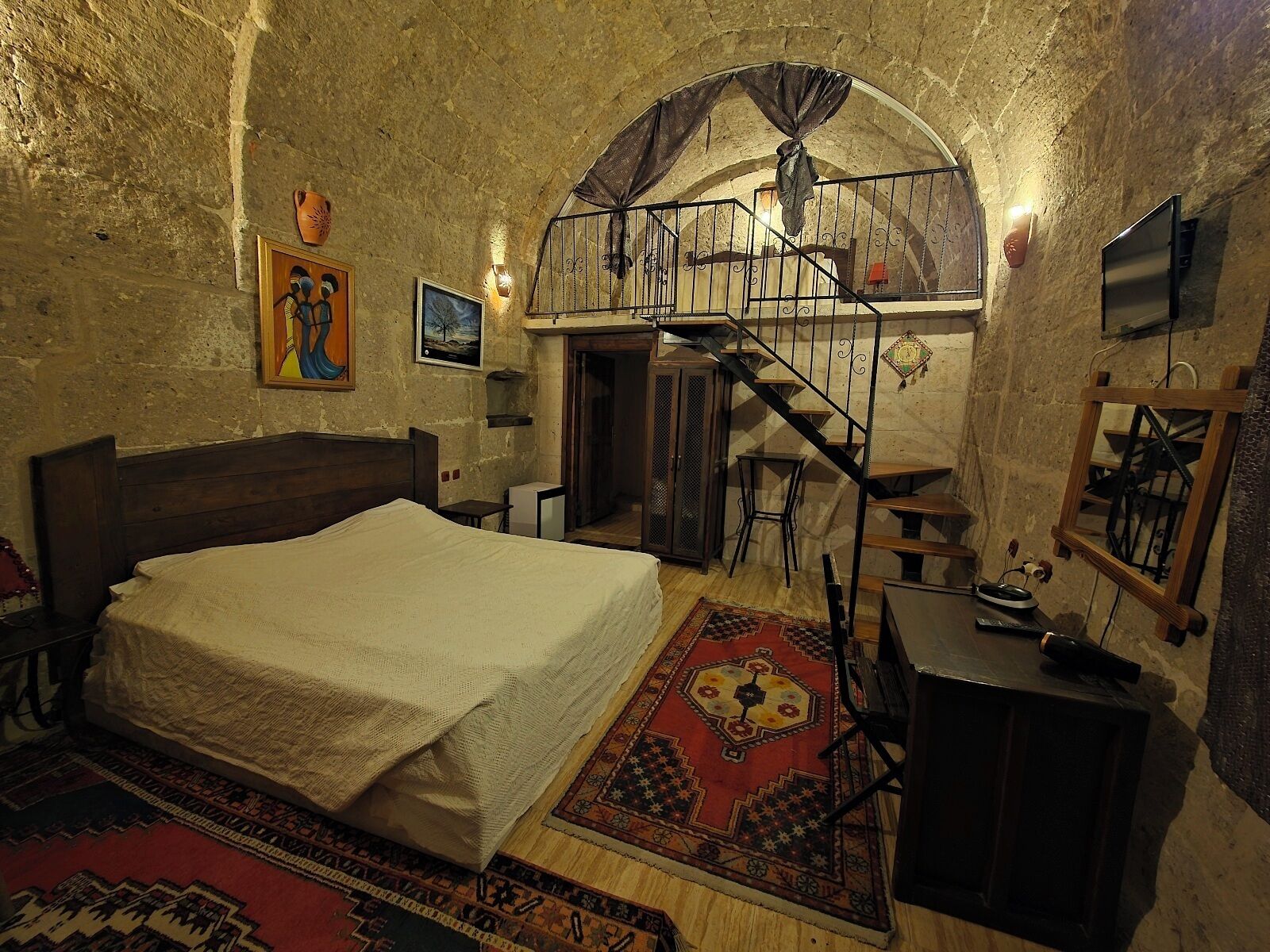 A bedroom with a bed, a desk, a chair, a TV, and a staircase leading to an upper level.
