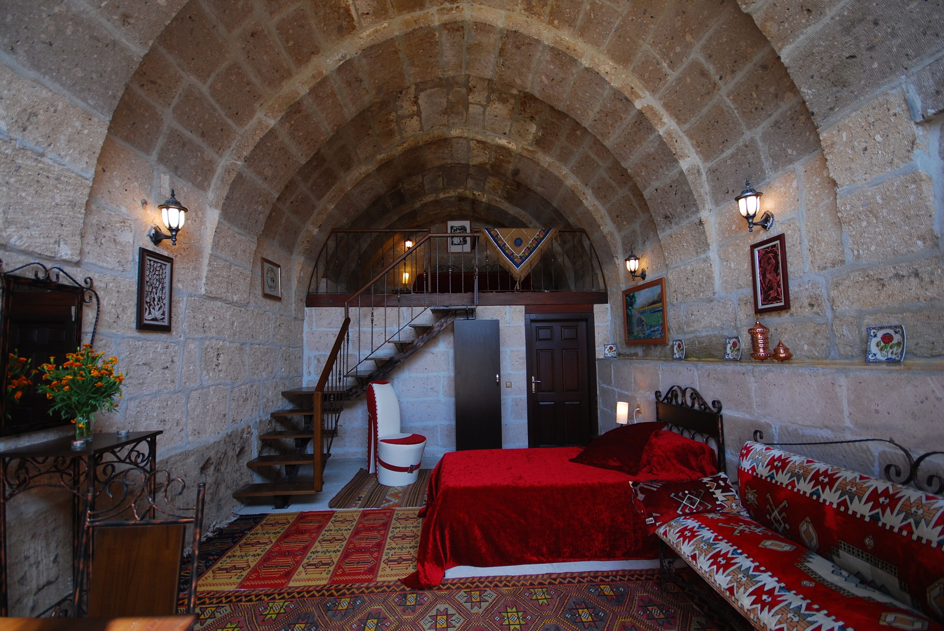 Una habitación con techo abovedado de piedra, una cama con colcha roja, un banco de madera, una escalera y una pequeña mesa con un jarrón de flores.