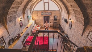 Una habitación con techo de piedra abovedado, una escalera de madera, una cama con manta roja y una pequeña mesa con lámpara.