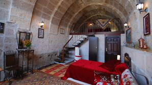 Una habitación con techo de piedra abovedado, una escalera, una cama con colcha roja y una mesita con un jarrón de flores.