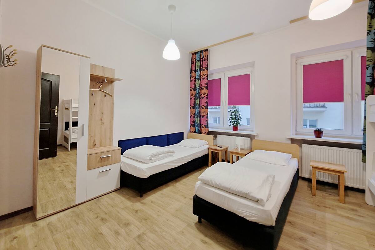 A modern hotel room with two beds, a wooden wardrobe, a mirror, a small table, and a window with pink blinds.