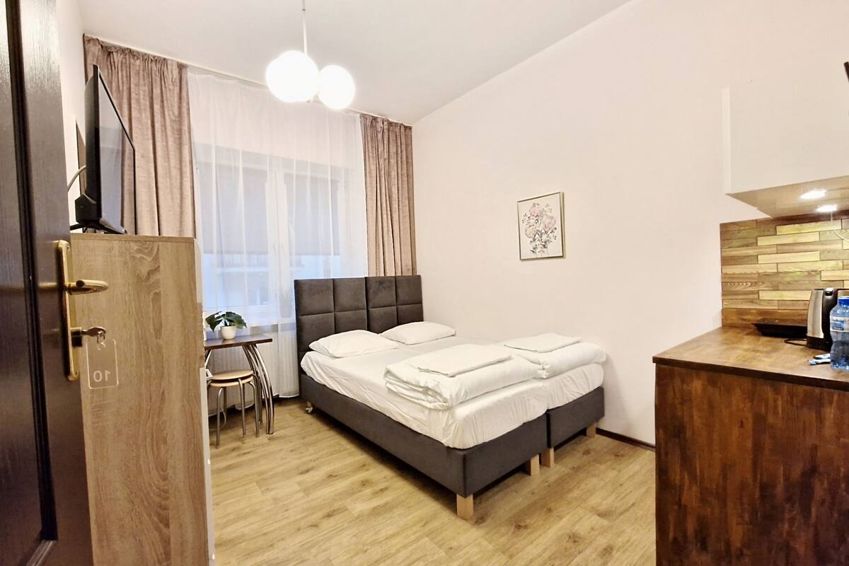 A hotel room with a bed, a desk, a chair, a small table, a window with curtains, a picture on the wall, and a wooden cabinet.