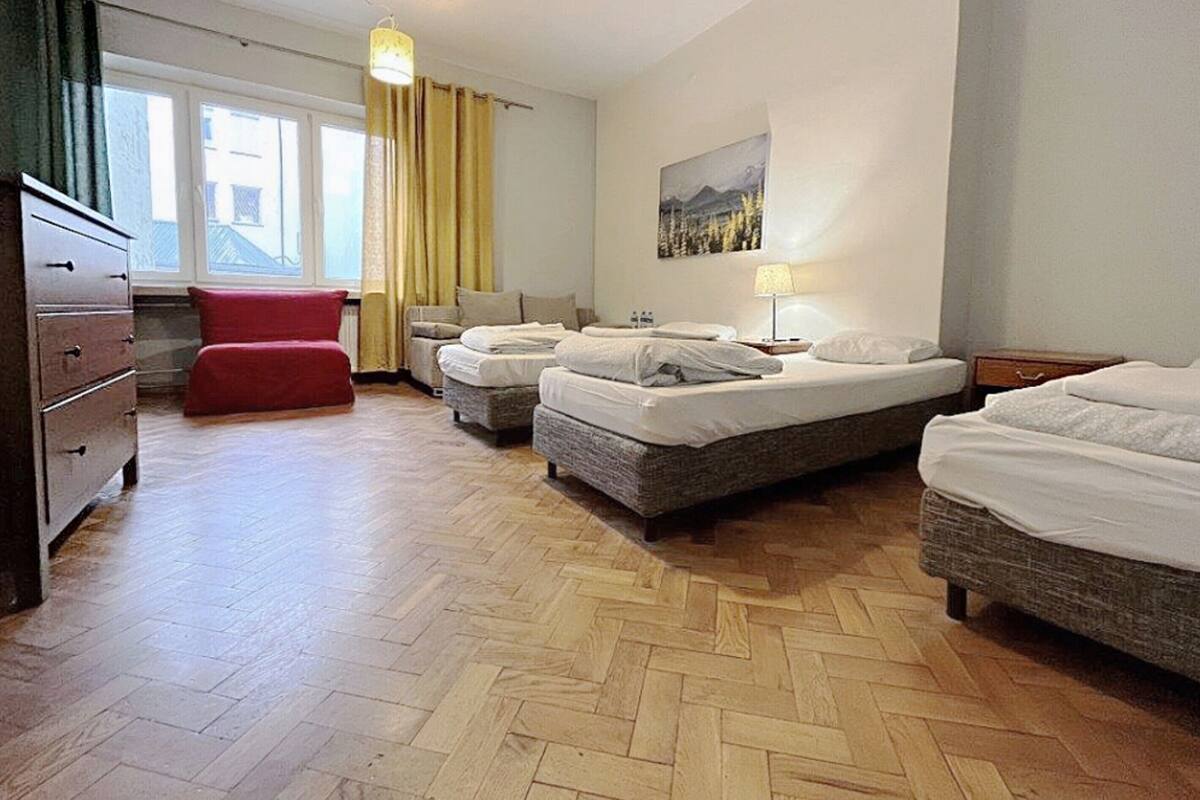 A hotel room with three beds, a wooden herringbone floor, a red chair, and a painting on the wall.