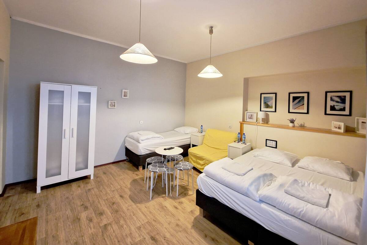 A hotel room with two beds, a wardrobe, a small table with chairs, a yellow chair, and a ceiling light.