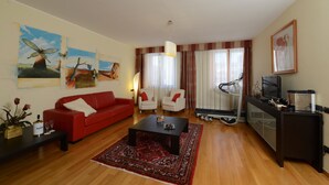 Una sala de estar con un sofá rojo, dos sillones blancos, una mesa de centro, una caminadora y un televisor sobre un mueble.