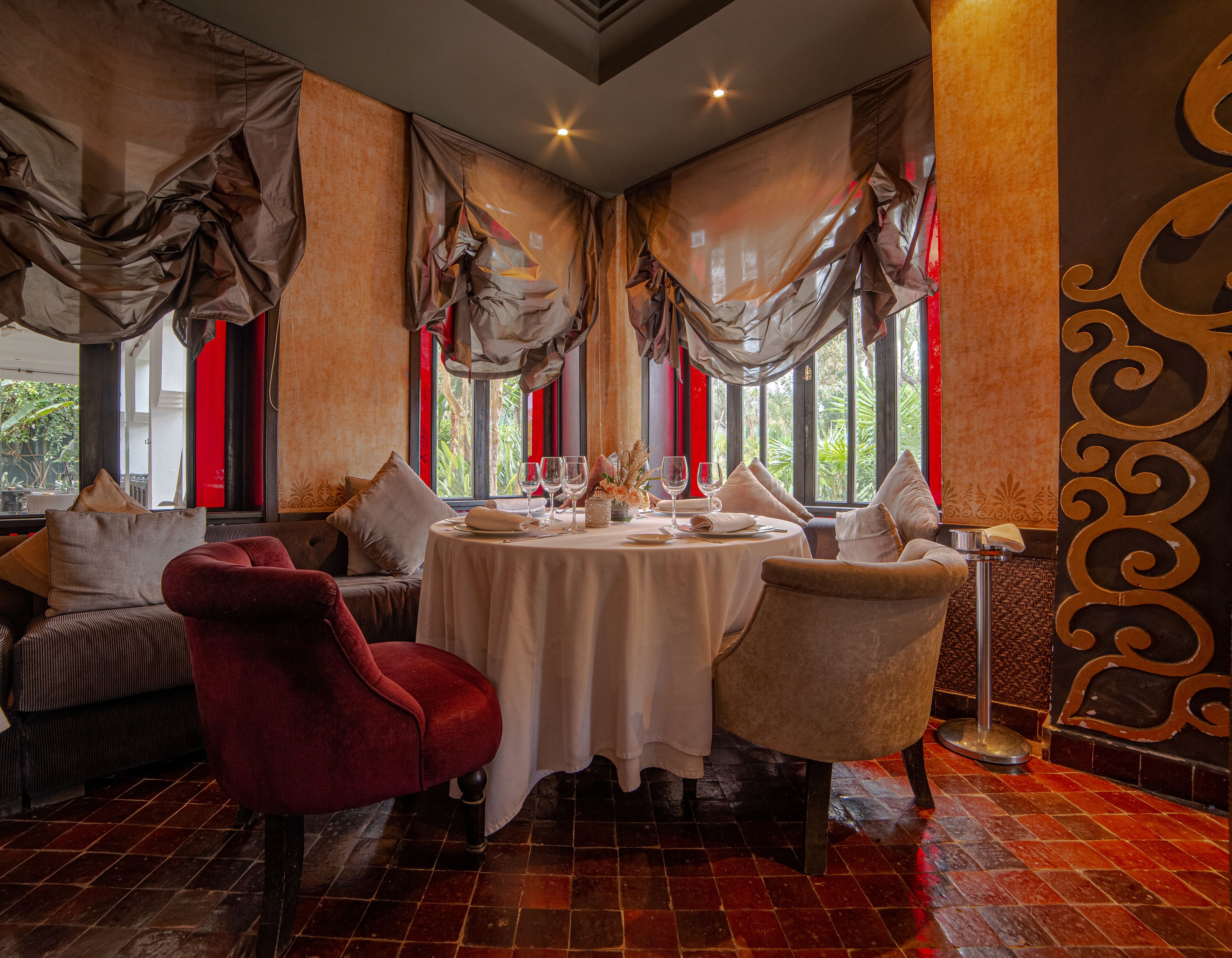A dining area with a round table set for two, surrounded by plush seating, large windows with red curtains, and a decorative wall with a golden pattern.