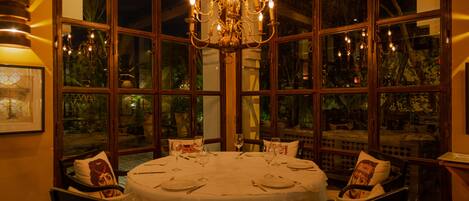 A dining room with a round table set for four, surrounded by chairs, large windows, and a chandelier.