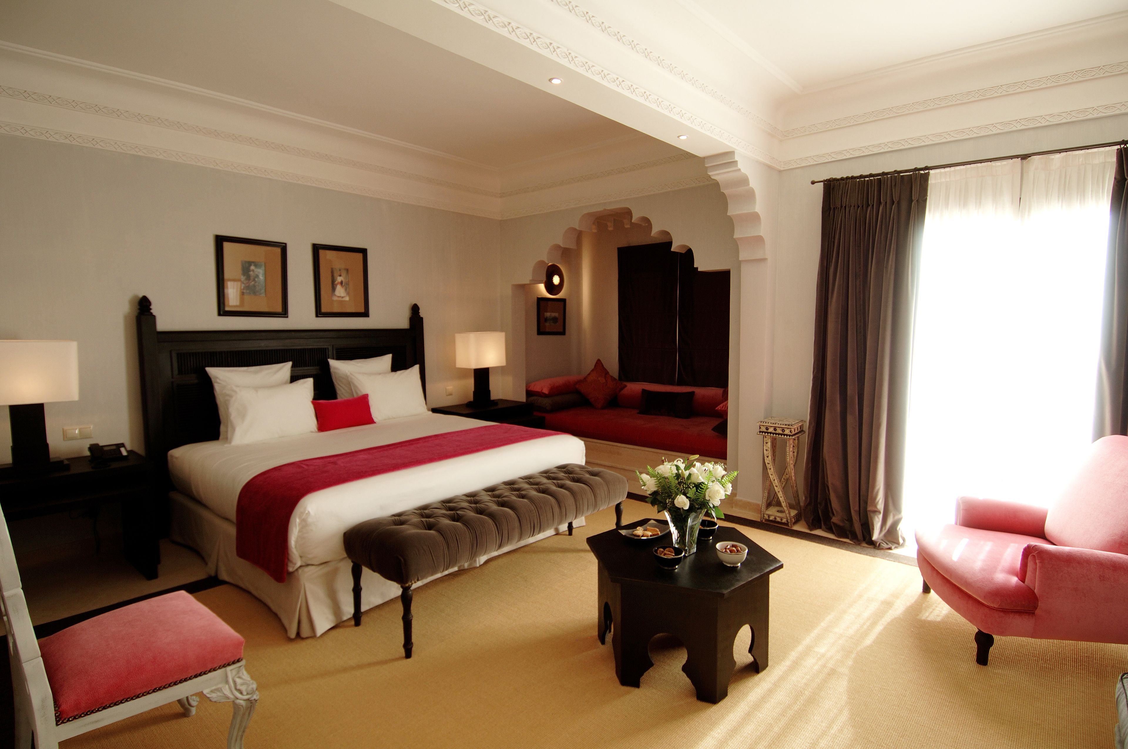 Habitación de hotel con una cama grande, un banco, una mesita con un jarrón, una silla y vistas al exterior a través de una ventana con cortinas.