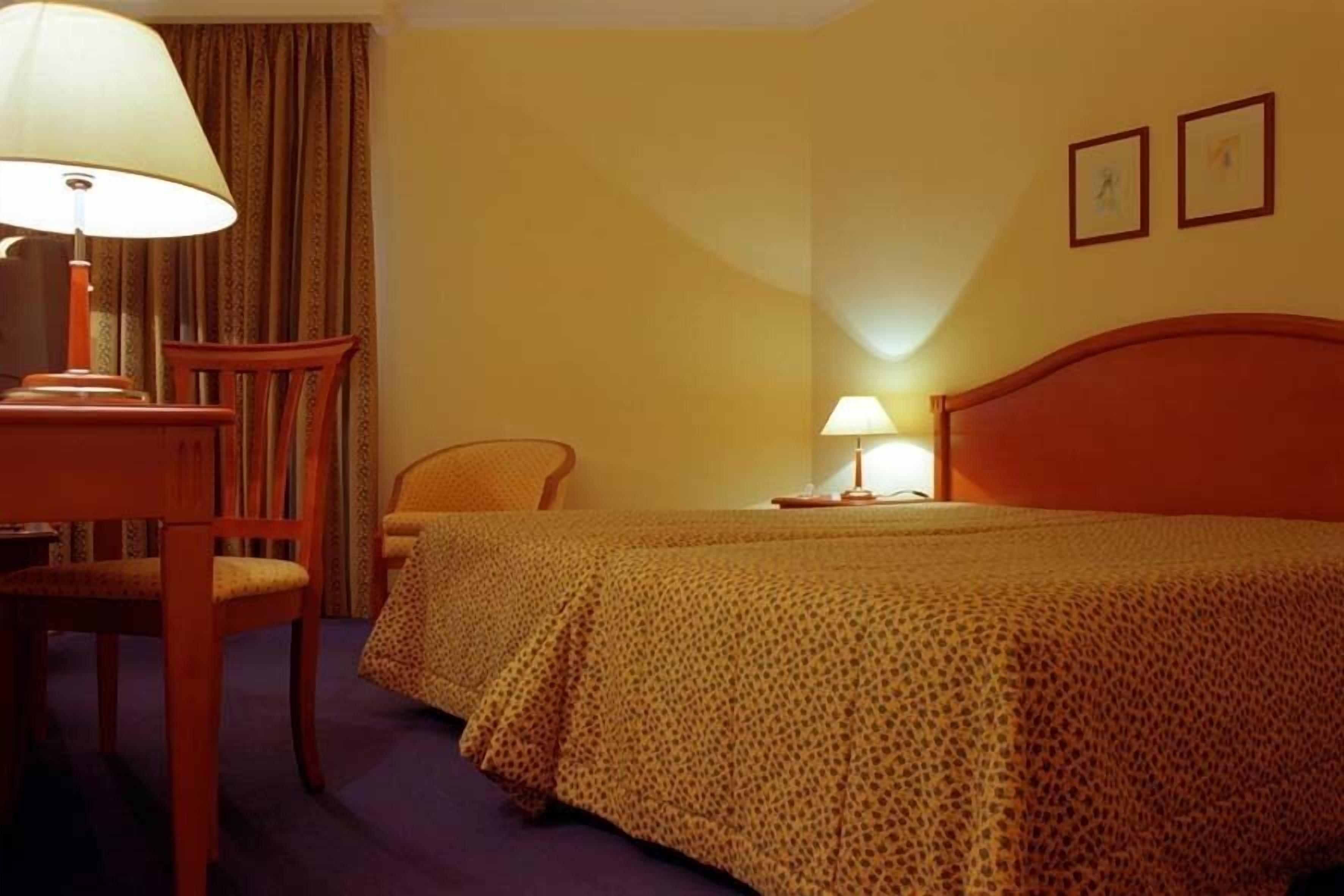 Quarto de hotel com uma cama, duas cadeiras, uma escrivaninha, um abajur e uma mesa de cabeceira.