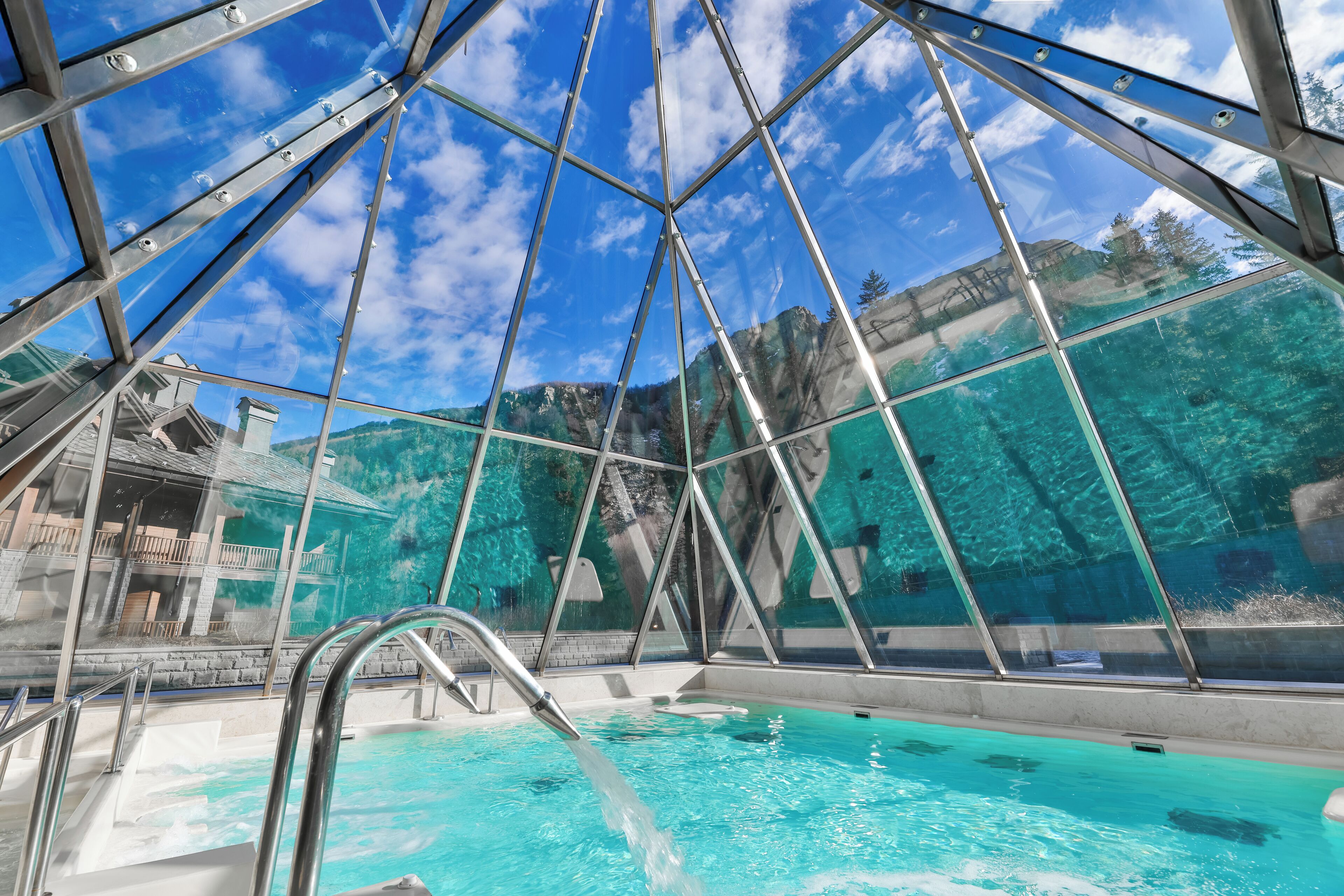 Una piscina interna con pareti di vetro trasparente, struttura ad intelaiatura metallica e vista su un paesaggio montuoso.